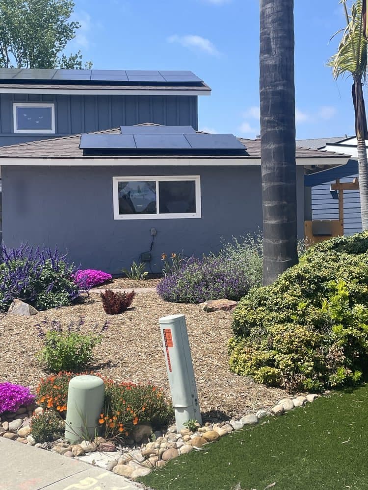 Black solar panels on the roof of a grey stucco home with a drought-tolerant front yard